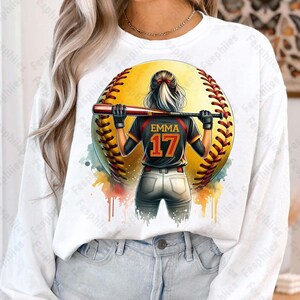 May include: A white long-sleeve shirt with a watercolor design of a softball player holding a bat. The player is wearing a black and red jersey with the number 17 and the name "EMMA" on the back. The softball is yellow with red stitching.