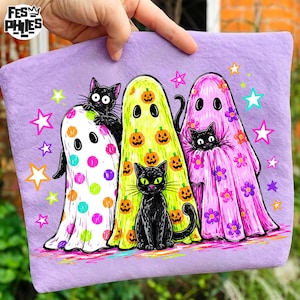 May include: A light purple fabric item featuring a graphic print of three ghosts and three black cats. The ghosts are white with colorful polka dots, yellow with orange pumpkins, and pink with purple flowers. Brightly colored stars surround the figures.