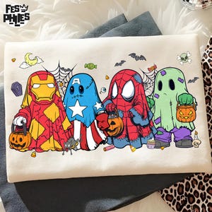 May include: A white t-shirt featuring a graphic of four characters dressed as ghosts for Halloween. The characters resemble Iron Man, Captain America, Spider-Man, and a green ghost, each holding a pumpkin candy bucket. The design includes bats, spiderwebs, and candy corn.