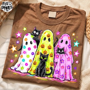 May include: A brown t-shirt featuring a graphic of three ghosts and two black cats. One ghost has colorful polka dots, one has orange pumpkins, and one has purple flowers. The design includes various colorful stars surrounding the figures.