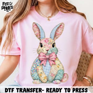 Puede incluir: Camiseta rosa claro con un diseño de conejo de retazos colorido. El conejo tiene orejas y secciones del cuerpo con estampado de cuadros azules y flores, y una pajarita rosa. El texto "DTF TRANSFER- READY TO PRESS" está en la parte inferior.