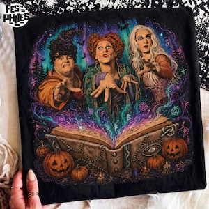 May include: A black t-shirt featuring a graphic print of three witches with hands outstretched over an open spellbook. The design includes swirling purple and teal magical energy, several orange jack-o'-lanterns, and a dark forest background.