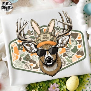 May include: White t-shirt featuring a deer wearing sunglasses and a camouflage hat with the text "egg Hunter". The deer has bunny ears and is surrounded by Easter eggs. The design is set against a green and orange background.