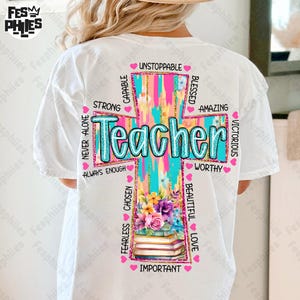 Op de afbeelding: Wit T-shirt met een kleurrijk kruisontwerp. Het kruis toont het woord "Teacher" in turquoise, omringd door inspirerende woorden en bloemen- en boekafbeeldingen. Het ontwerp bevat roze, turquoise en gele kleuren.