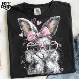 Puede incluir: Camiseta negra con una ilustraci&oacute;n de un conejito con gafas rosas en forma de coraz&oacute;n y un lazo rosa. El dise&ntilde;o incluye corazones y flores rosas. La camiseta es de material suave.