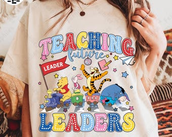 Puede incluir: Camiseta color crema con un gráfico colorido que dice "TEACHING future LEADERS". El diseño incluye ilustraciones de Winnie the Pooh, Piglet, Tigger y Eeyore en un tren, con estrellas y una bandera "LEADER".