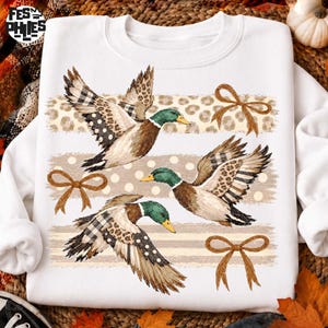May include: A white crewneck sweatshirt featuring a graphic print of three flying mallard ducks. The ducks have green heads and wings patterned with leopard print, plaid, and polka dots. Three brown ribbon bows are placed between the ducks.
