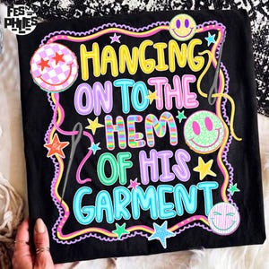 May include: A black t-shirt featuring a colorful graphic with the text HANGING ON TO THE HEM OF HIS GARMENT. The design includes yellow, purple, and blue lettering, multiple smiley faces with various patterns, sewing needles, and small stars.