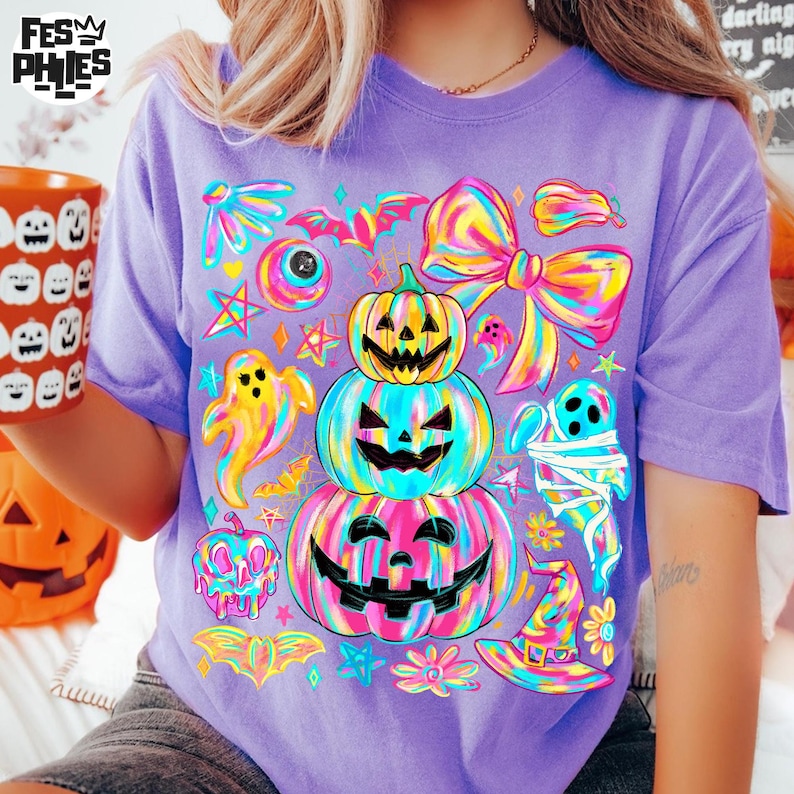 May include: A purple t-shirt featuring a colorful Halloween-themed design. The design includes stacked jack-o'-lanterns, ghosts, bats, and a witch's hat. The shirt has a relaxed fit and short sleeves.