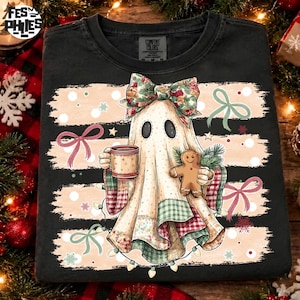 May include: A black t-shirt featuring a graphic of a ghost wearing a floral bow, holding a mug and a gingerbread man. The ghost is draped in a patchwork blanket with plaid patterns, surrounded by light pink horizontal stripes and bows.