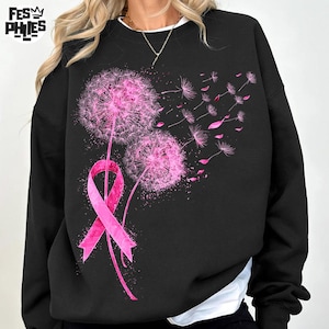May include: Black sweatshirt featuring a pink ribbon and two pink dandelion flowers with seeds blowing away. The design is a graphic print. The sweatshirt has a crew neck and long sleeves.