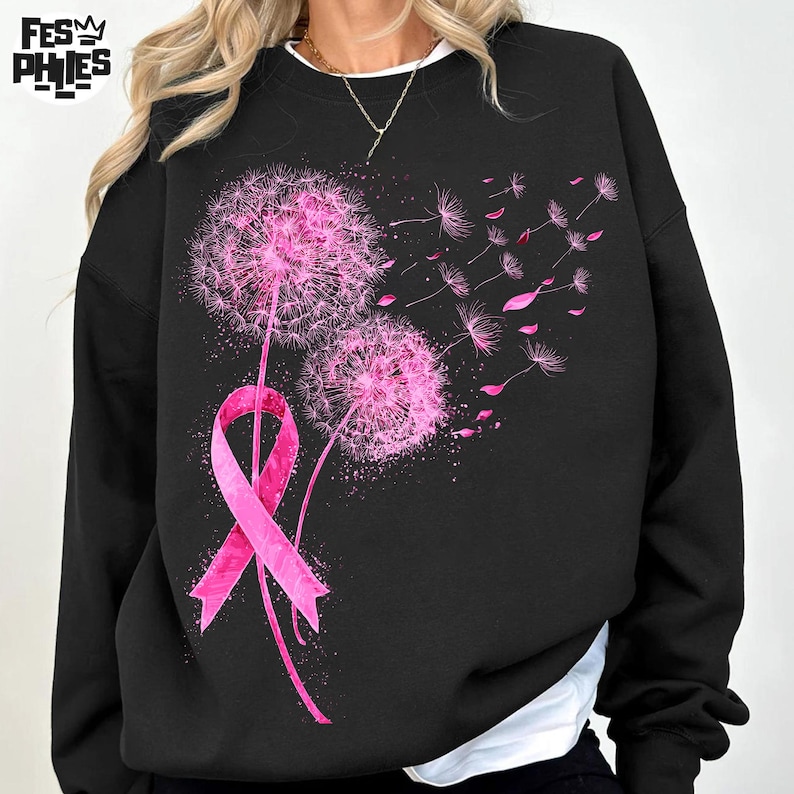 May include: Black sweatshirt featuring a pink ribbon and two pink dandelion flowers with seeds blowing away. The design is a graphic print. The sweatshirt has a crew neck and long sleeves.