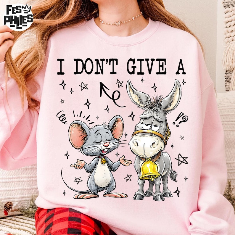 May include: Pink sweatshirt with the text "I DON'T GIVE A" above a cartoon mouse and donkey. The mouse is gray with pink ears, and the donkey has a gold bell. The design includes stars and other graphic elements.