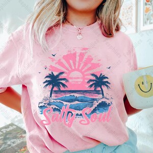 May include: Pink t-shirt featuring a graphic of a sunset over the ocean with palm trees and the text "Salty Soul".
