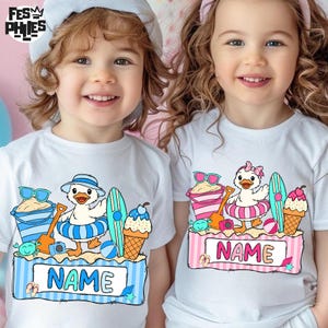 May include: Two white t-shirts with a colorful beach-themed graphic. Each shirt features a cartoon duck in a swim ring, sunglasses, a surfboard, and an ice cream cone. The shirts have a blue and pink striped border with the word "NAME".