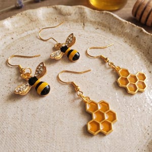 May include: Gold-toned bee and honeycomb earrings. Two pairs of earrings feature bee designs with black and yellow striped bodies and rhinestone wings. The other two earrings have a honeycomb design with yellow enamel.