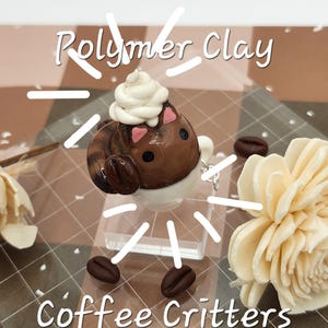 May include: A polymer clay figurine of a cat in a coffee cup, with whipped cream on its head. The figurine is brown with pink accents and sits on a clear stand, surrounded by coffee beans and a decorative flower. The text "Coffee Critters" is visible.