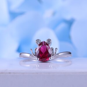 May include: A silver ring featuring a frog design. The ring has a pear-shaped, deep red gemstone as the frog's body, with two small, round, clear gemstones for eyes. The ring's band and frog details are silver.