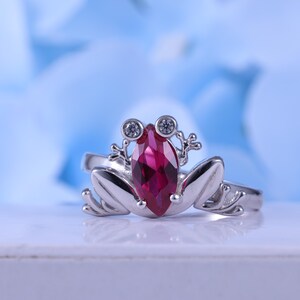 May include: A silver frog ring with a marquise-cut, deep red gemstone as the frog's body. The ring features two small, round, clear gemstones for eyes and detailed frog feet. The background is a soft blue.