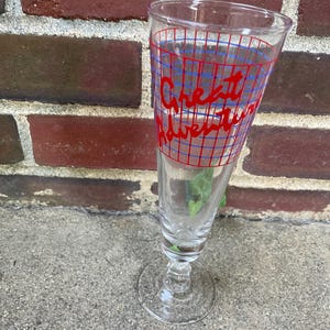 May include: A clear glass with a stem, featuring the words "Great Adventure" in red script, overlaid on a blue grid pattern. The glass is empty and stands on a concrete surface, with a brick wall in the background.