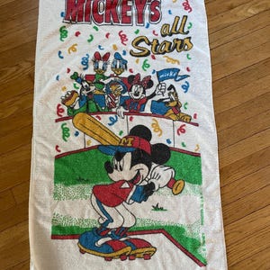 May include: A white rectangular beach towel featuring a colorful graphic of Mickey Mouse playing baseball with other Disney characters. The text reads Mickey's all Stars. The design includes confetti, a green baseball field, and red accents.