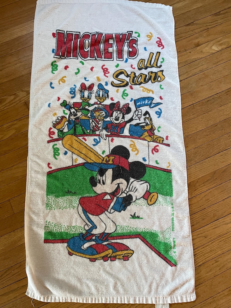 May include: A white rectangular beach towel featuring a colorful graphic of Mickey Mouse playing baseball with other Disney characters. The text reads Mickey's all Stars. The design includes confetti, a green baseball field, and red accents.