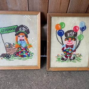 May include: Two framed embroidered pictures. One features a girl with orange braids and a lemonade stand, the sign reads "LEMONADE". The other shows a clown holding balloons. Both have wooden frames and are on a beige background.