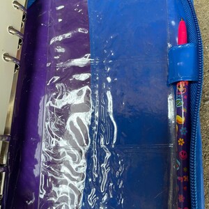 Puede incluir: Un archivador azul con cremallera y un interior morado y azul. El archivador contiene fundas de pl&aacute;stico transparente y un bol&iacute;grafo con punta rosa. El bol&iacute;grafo est&aacute; decorado con flores de colores y el n&uacute;mero 24.