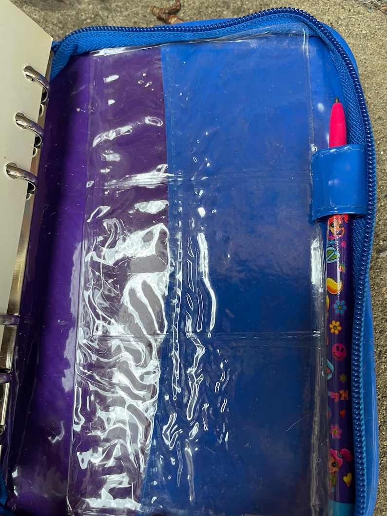 Puede incluir: Un archivador azul con cremallera y un interior morado y azul. El archivador contiene fundas de pl&aacute;stico transparente y un bol&iacute;grafo con punta rosa. El bol&iacute;grafo est&aacute; decorado con flores de colores y el n&uacute;mero 24.