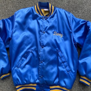 May include: A blue satin jacket with the name "Danny" embroidered in gold script. The jacket has a button-up front, pockets, and yellow and blue striped cuffs and collar. A vintage style jacket.