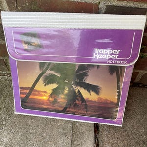 May include: A purple Trapper Keeper notebook with a white textured top flap. The front cover features a sunset scene over the ocean with palm trees in silhouette. The text Trapper Keeper NOTEBOOK is printed in white on the upper right side.