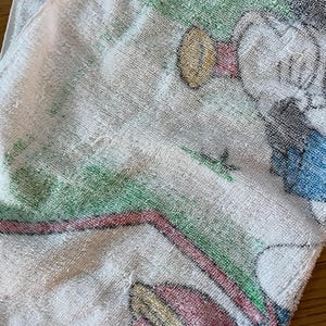 May include: A white terry cloth towel featuring a printed graphic of a cartoon character with a red hat, blue clothing, and yellow shoes. The design includes green grassy areas and a red curved line, rendered in a soft, textured fabric finish.