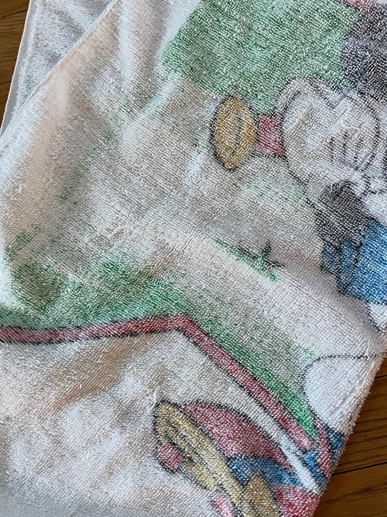 May include: A white terry cloth towel featuring a printed graphic of a cartoon character with a red hat, blue clothing, and yellow shoes. The design includes green grassy areas and a red curved line, rendered in a soft, textured fabric finish.
