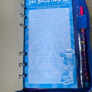 Puede incluir: Carpeta azul con cremallera con papel rayado y un bol&iacute;grafo. El papel tiene el texto "you gotta have it!" y "Cool!" con un personaje de dibujos animados. El bol&iacute;grafo es morado con un dise&ntilde;o decorativo.