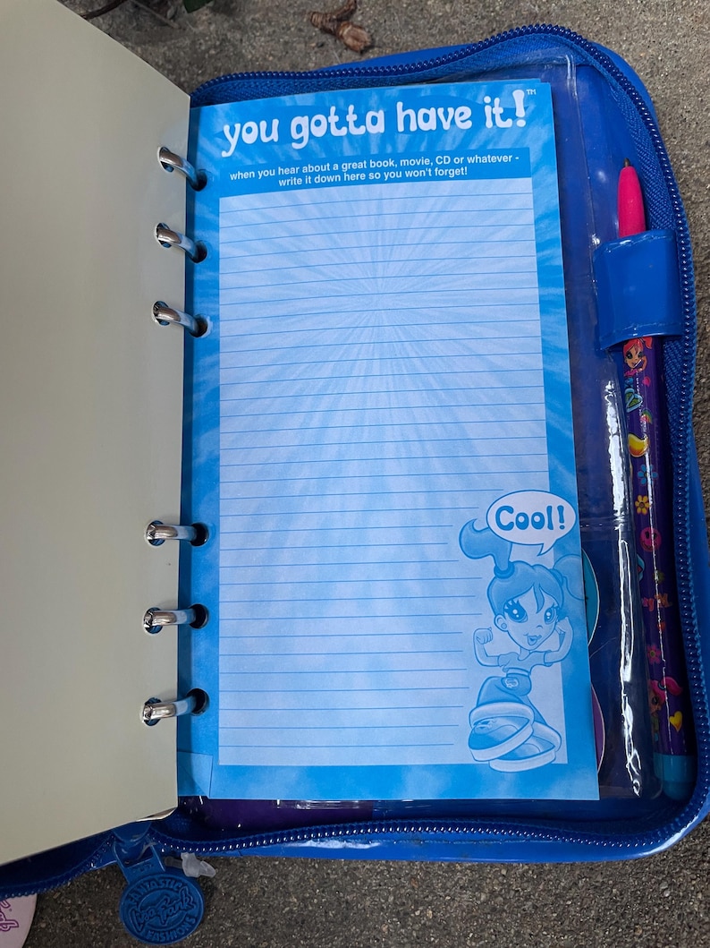 Puede incluir: Carpeta azul con cremallera con papel rayado y un bol&iacute;grafo. El papel tiene el texto "you gotta have it!" y "Cool!" con un personaje de dibujos animados. El bol&iacute;grafo es morado con un dise&ntilde;o decorativo.