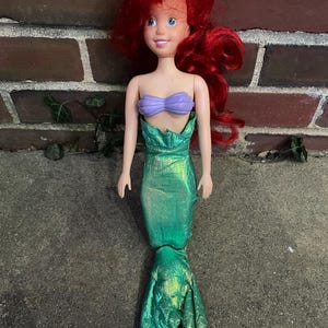 May include: A doll of a mermaid with red hair, a purple top, and a green, shimmering tail. The doll is standing upright, with arms at its sides. The doll's face has a smiling expression.