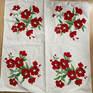 May include: A white rectangular fabric cloth featuring four repeating floral clusters. Each cluster consists of vibrant red flowers with yellow centers and green leaves, arranged in a symmetrical grid pattern across the surface of the textile.