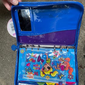 Puede incluir: Carpeta de vinilo azul abierta con un bolsillo transparente, espejo y forro interior morado. En el interior hay pegatinas coloridas, un bol&iacute;grafo y un calendario. La carpeta presenta el texto "Girls Rule" y gr&aacute;ficos de dibujos animados.