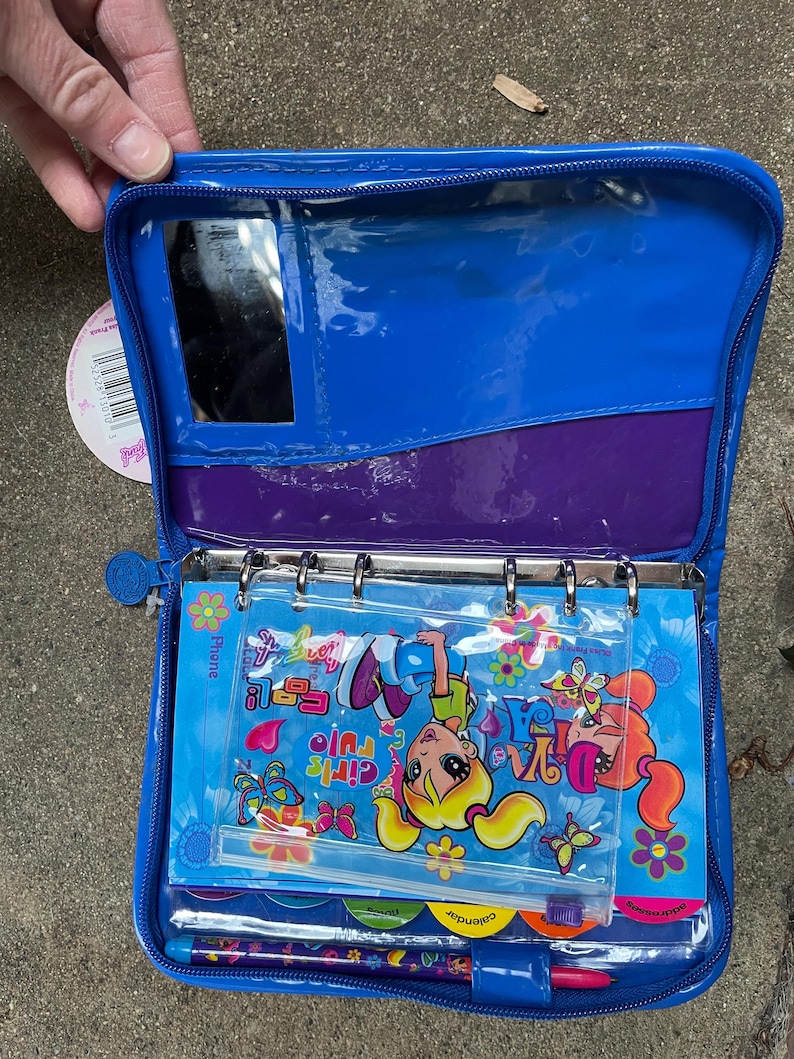 Puede incluir: Carpeta de vinilo azul abierta con un bolsillo transparente, espejo y forro interior morado. En el interior hay pegatinas coloridas, un bol&iacute;grafo y un calendario. La carpeta presenta el texto "Girls Rule" y gr&aacute;ficos de dibujos animados.