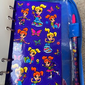 Puede incluir: Un archivador azul con cremallera con una hoja de pegatinas con dibujos de chicas y mariposas. Las pegatinas est&aacute;n sobre un fondo morado. Un bol&iacute;grafo rosa con un dise&ntilde;o floral est&aacute; en un soporte en el lado derecho del archivador.