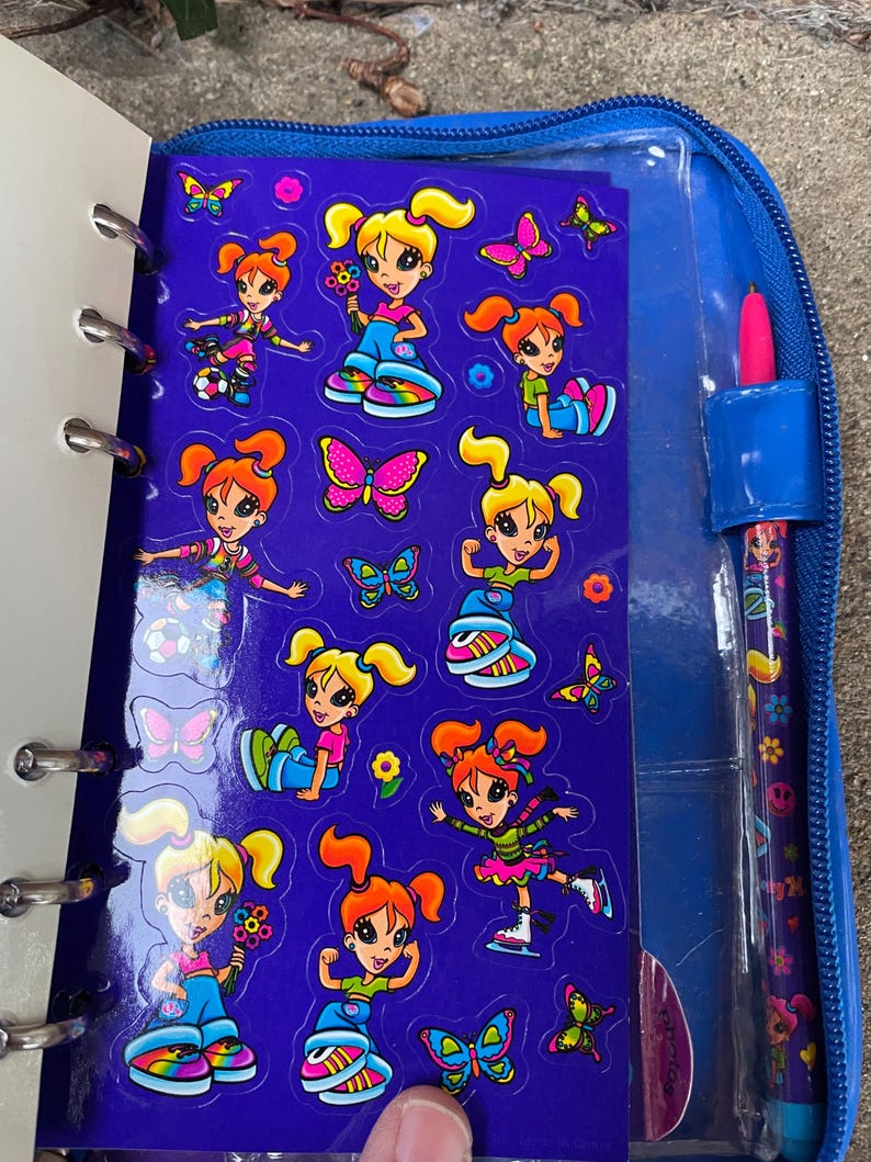 Puede incluir: Un archivador azul con cremallera con una hoja de pegatinas con dibujos de chicas y mariposas. Las pegatinas est&aacute;n sobre un fondo morado. Un bol&iacute;grafo rosa con un dise&ntilde;o floral est&aacute; en un soporte en el lado derecho del archivador.