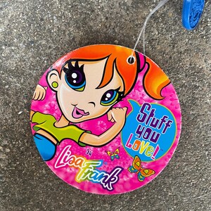 Puede incluir: Una etiqueta redonda de Lisa Frank con una ilustraci&oacute;n de una chica de dibujos animados. La etiqueta es rosa con el texto "Stuff You Love!" en una burbuja azul. El logotipo de Lisa Frank y gr&aacute;ficos de mariposas tambi&eacute;n son visibles.