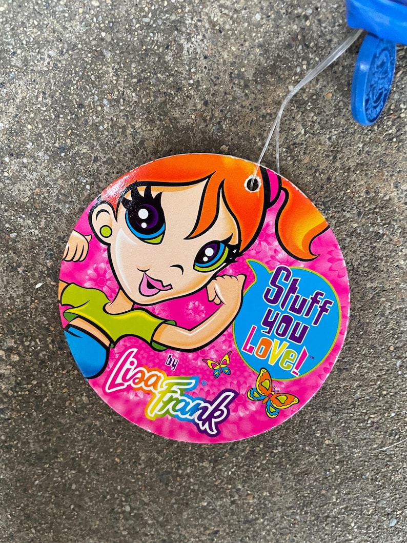 Puede incluir: Una etiqueta redonda de Lisa Frank con una ilustraci&oacute;n de una chica de dibujos animados. La etiqueta es rosa con el texto "Stuff You Love!" en una burbuja azul. El logotipo de Lisa Frank y gr&aacute;ficos de mariposas tambi&eacute;n son visibles.