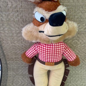 May include: A plush Wile E. Coyote doll. The coyote has a brown and tan fur pattern, large eyes, and a black nose. He wears a red and white checkered shirt, tan pants, and a brown belt that says "Wile E. Coyote".