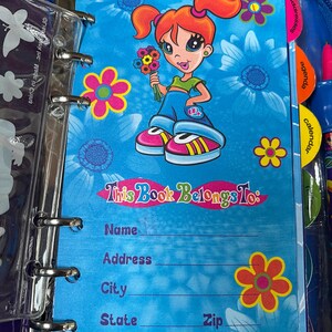Puede incluir: Una carpeta azul con un dise&ntilde;o floral y una ilustraci&oacute;n de una ni&ntilde;a de dibujos animados. La p&aacute;gina dice "This Book Belongs To:" seguida de l&iacute;neas para nombre, direcci&oacute;n, ciudad, estado, c&oacute;digo postal y tel&eacute;fono. La carpeta tiene pesta&ntilde;as para direcciones, agenda y calendario.
