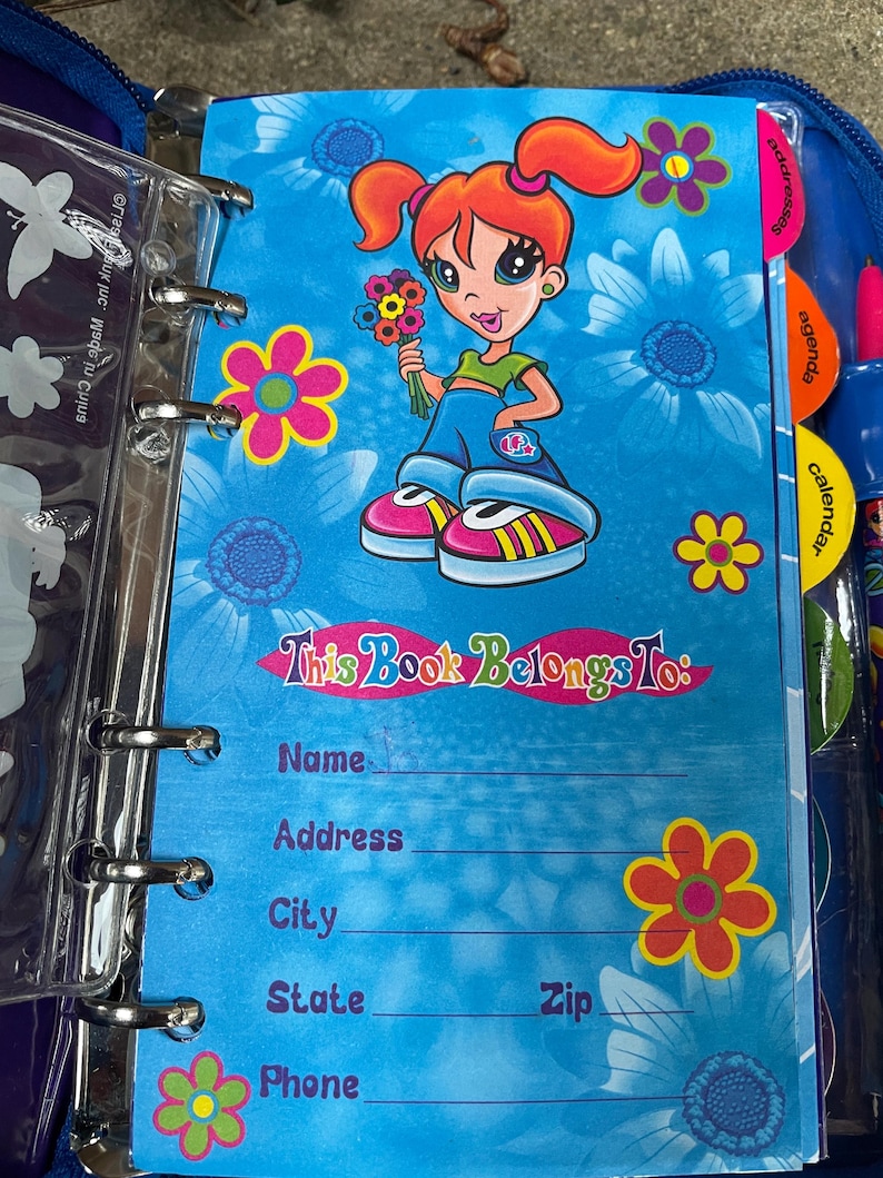 Puede incluir: Una carpeta azul con un dise&ntilde;o floral y una ilustraci&oacute;n de una ni&ntilde;a de dibujos animados. La p&aacute;gina dice "This Book Belongs To:" seguida de l&iacute;neas para nombre, direcci&oacute;n, ciudad, estado, c&oacute;digo postal y tel&eacute;fono. La carpeta tiene pesta&ntilde;as para direcciones, agenda y calendario.