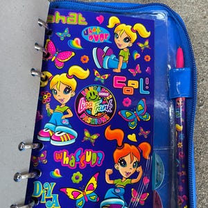 Puede incluir: Un cuaderno azul con tem&aacute;tica de Lisa Frank y un bol&iacute;grafo a juego. El cuaderno presenta personajes de dibujos animados, mariposas y frases como "What's Up?" y "Sweet!". El bol&iacute;grafo y el cuaderno est&aacute;n en un estuche azul con cremallera.
