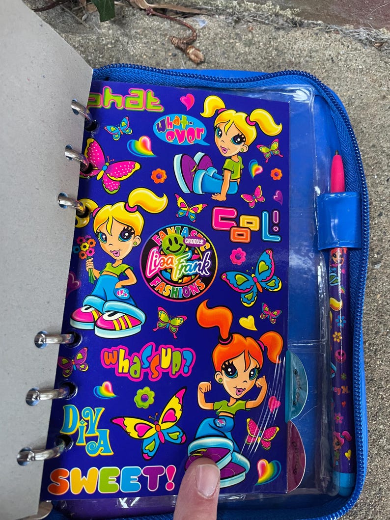 Puede incluir: Un cuaderno azul con tem&aacute;tica de Lisa Frank y un bol&iacute;grafo a juego. El cuaderno presenta personajes de dibujos animados, mariposas y frases como "What's Up?" y "Sweet!". El bol&iacute;grafo y el cuaderno est&aacute;n en un estuche azul con cremallera.
