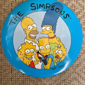 May include: A round, blue button featuring the Simpsons family. The image includes Homer, Marge, Bart, Lisa, and Maggie. The text "THE SIMPSONS" is printed around the edge of the button. The artist's signature, "Matt Groening," is visible.