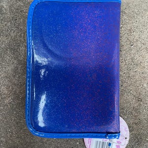 Puede incluir: Un estuche azul brillante con un borde azul a juego. El estuche tiene un acabado brillante y un cierre de cremallera. Una etiqueta redonda con un c&oacute;digo de barras est&aacute; adjunta en la esquina inferior derecha.